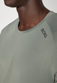 Olivno zelena športna majica z gladko teksturo, okroglo ovratnico in črnimi "BORG" logotipom na rami. Vidni so šivi.
