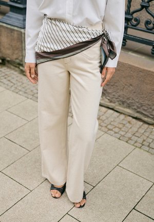 Personne portant un pantalon beige à jambes larges, une chemise blanche boutonnée, des sandales noires ouvertes à l’avant, et un foulard en soie rayé noué à la taille.
