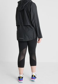 Chaqueta ligera negra con capucha, combinada con leggings negros que tienen paneles de malla y zapatillas deportivas con una suela colorida.