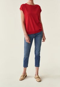 Rotes kurzärmliges Oberteil, lockere Passform, Strickstoff. Kombiniert mit hellblauen knöchellangen Jeans und beigen Slippern mit goldenen Elementen.