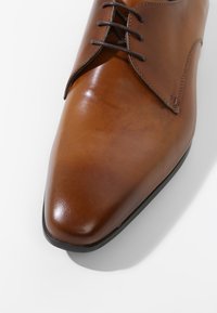 Chaussure habillée en cuir marron avec un bout pointu, texture lisse et coutures minimales. Présente des lacets marron et une semelle noire.