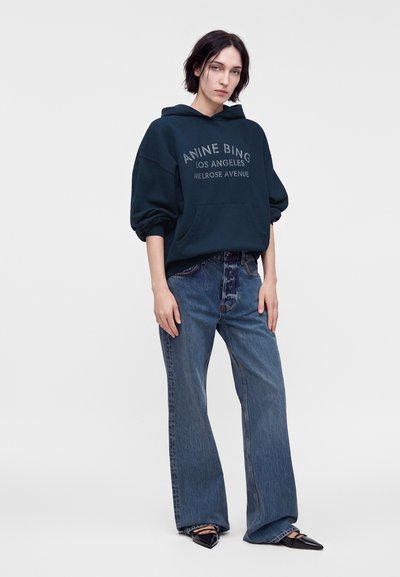 Νεαρό άτομο που φοράει σκούφο oversized φούτερ με γράμματα, άνετο μπλε τζιν και μαύρα μυτερά παπούτσια, στέκεται μπροστά σε ένα απλό φόντο.