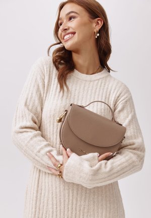 Hellbraune Lederhandtasche mit einer runden Klappe und einem oberen Tragegriff. Das Modell trägt einen cremefarbenen, gerippten Pullover, der die Textur und Farbe der Tasche zur Geltung bringt.