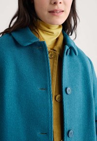 Femme portant un col roulé superposé jaune moutarde et un cardigan sous un manteau bleu texturé à boutons, vue de la mâchoire au torse.