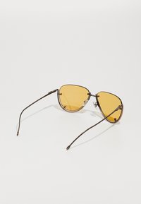Occhiali da sole a forma di occhio di gatto con lenti gialle, montatura in metallo e sottili stanghette marroni. I cuscinetti nasali regolabili migliorano comfort e vestibilità.