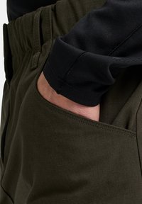 Olivgrüne Hose aus strukturiertem Stoff, mit einer Seitentasche und einem elastischen Bund. Darunter ein schwarzes Langarmshirt.