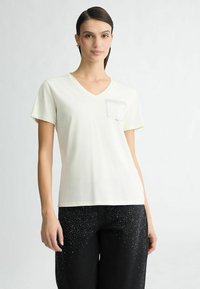 Cremefarvet bomuld t-shirt med V-hals, korte ærmer og en lomme på venstre side, prydet med rhinsten. Båret med sorte bukser med juveler.