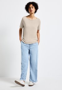 Beige kortärmad topp med vid halsringning, matchad med ljusblå vida jeans och vita sneakers. Mjuk tygkvalitet, avslappnad passform, avslappnad stil.