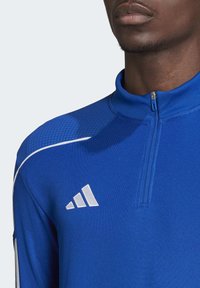 Maglietta atletica blu con zip a metà, con parte superiore testurizzata, accenti bianchi e logo Adidas sul lato sinistro del petto. Il materiale sembra liscio e traspirante.