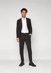 Costume noir à coupe ajustée, associé à une chemise blanche à col boutonné et des chaussures noires. Présente un revers classique, une texture lisse et un design sur mesure.