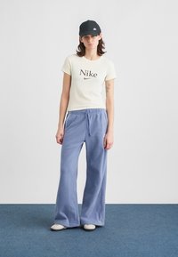 Ljusgul Nike-t-shirt med brunt logotyp, kombinerad med lösa, ljusblå joggers. Modellen bär en svart keps och enkla grå skor.