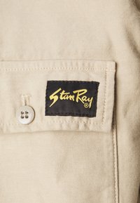 Beige fabric shirt with a black label featuring yellow "Stan Ray" text. Includes a silver button and stitched details.