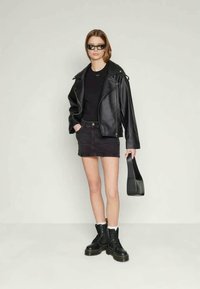 Veste en cuir noire sur un t-shirt noir, associée à une jupe en denim noire. Bottes de combat noires et lunettes de soleil, tenant un sac à main noir.