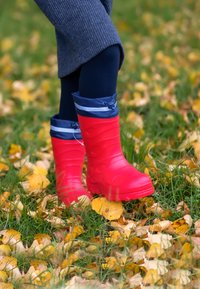 Rode rubberen regenlaarzen met een blauwe, opvouwbare manchet en trekkoorddetail, staand in het gras tussen gele herfstbladeren.