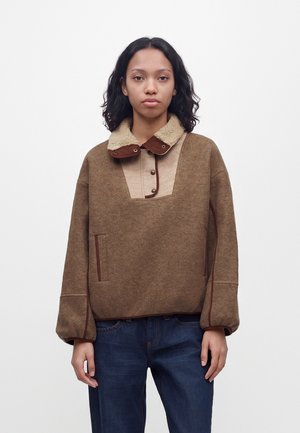 Une femme porte un pull marron à col en polaire crème et avec une poche à l’avant, associé à un jean bleu foncé, sur un fond blanc uni.