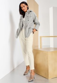 Hellgraue Zip-Jacke mit Kapuze, kombiniert mit cremfarbenen Hosen. Die Schuhe verfügen über spitze, schlangenledergemusterte Absätze. Minimalistisches Design.