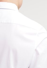 Camicia bianca da uomo con una texture liscia, dotata di un colletto classico e un design pulito e minimalista, vista da dietro.