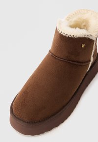 Bottine marron en daim avec une doublure en polaire crème douce. Présente des accents cousus et une semelle en caoutchouc. Design simple et confortable.