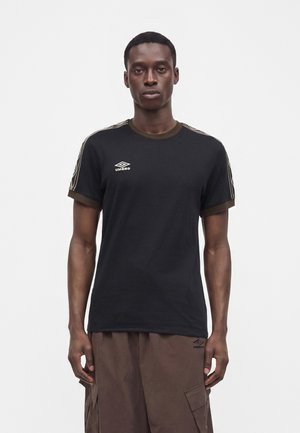 Νέος άνδρας με μαύρη μπλούζα Umbro με καφέ διακόσμηση και καφέ παντελόνι cargo, στέκεται μπροστά σε απλό λευκό φόντο.