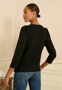 Fekete felső 3/4-es ujjal, kerek nyakkivágással és ráncolt vállrészletekkel, kék farmernadrággal párosítva. Sima textúra, minimalista dizájn.