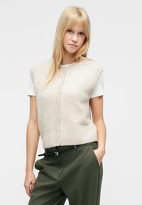 Bézs rövid ujjú kötött pulóver kerek nyakkal és gombos záródással, olívazöld magas derekú nadrággal és fekete övvel párosítva.