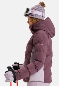 Puffige lila und hellgraue Skijacke mit Kapuze, ausgestattet mit Handschuhen und Skistöcken, mit Roxy-Logo und strukturiertem Strickmütze.