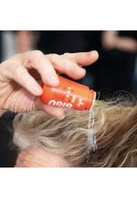 Flacon de produit capillaire orange incliné pour verser du liquide sur des cheveux ondulés et clairs, avec quelques gouttes visibles en suspension dans l'air.