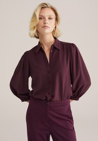 Chemisier coupe boutonnée bordeaux en tissu léger, doté d'un col et de manches longues bouffantes. Porté avec un pantalon bordeaux assorti.