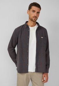 Marineblokjes overhemd met rode en witte ruiten, knoopsluiting en een zak op de linkerborst; gedragen over een wit T-shirt.