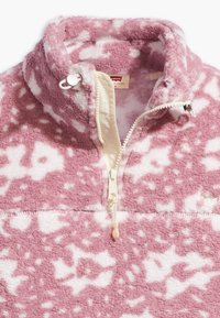 Różowy polarowy sweter z białym abstrakcyjnym wzorem, zamek błyskawiczny do połowy długości z kremową zakładką, wysoki kołnierz i miękka tekstura.