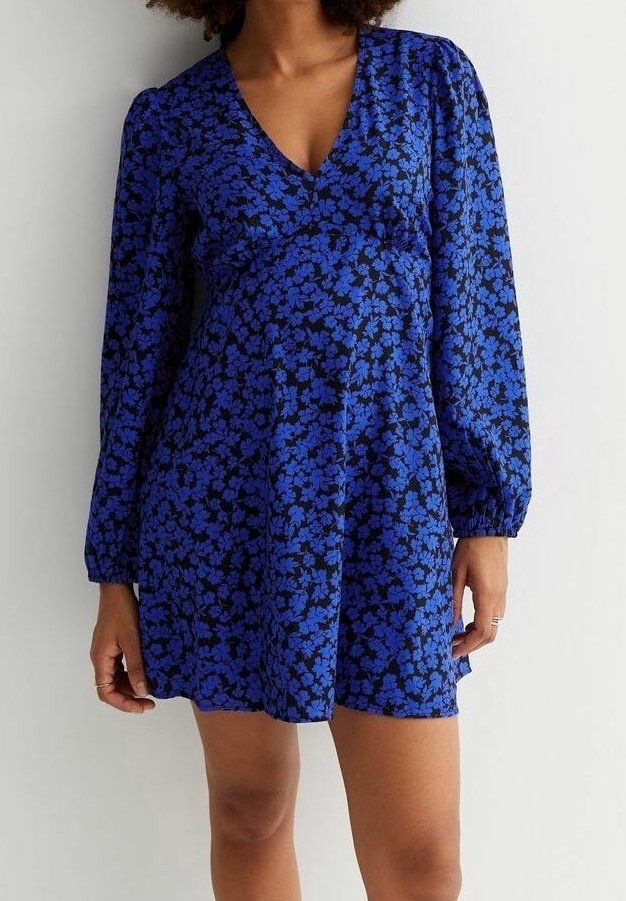 Femme portant une robe courte à manches longues, bleue à motifs floraux, avec un col en V et une jupe légèrement évasée, sur un fond clair uni.
