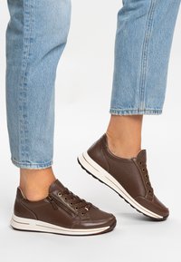 Brown leather sneakers feature a rounded shape, lacing system, and side zipper. Contrasting white sole with textured pattern and grip.