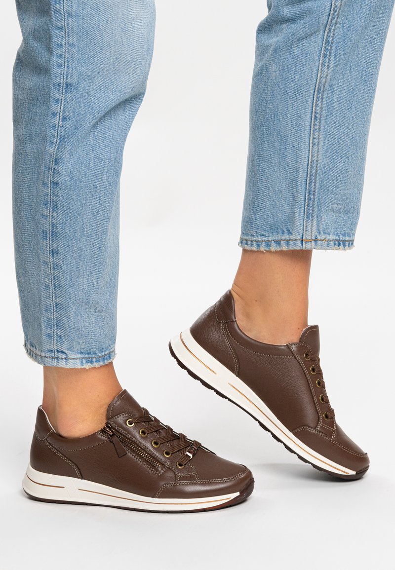 Brown leather sneakers feature a rounded shape, lacing system, and side zipper. Contrasting white sole with textured pattern and grip.