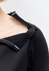 Vêtement noir avec une épaule ouverte maintenue par une large bande Velcro avec des surpiqûres blanches sur le haut de la poitrine et de l'épaule d'une personne.
