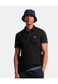Svart polo skjorta med en gul logotyp, kombinerad med en marinblå bucket cap. Modellen bär en blå undertröja och har en svart axelrem.