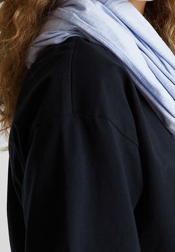 Personne portant un vêtement noir avec un tissu bleu clair à texture douce drapé autour du cou et de longs cheveux bruns bouclés.