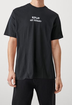 Mand iført en sort t-shirt med hvid tekst "REPLAY NOT ORDINARY" centreret på brystet, kombineret med mørke bukser mod en ensfarvet baggrund.