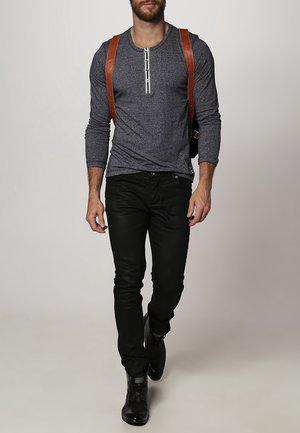 Dunkelgraues Langarmshirt mit knöpfbarem Ausschnitt, kombiniert mit schwarzen Jeans und schwarzen Stiefeln. Braune Leder-Tragegurte runden das Outfit ab.