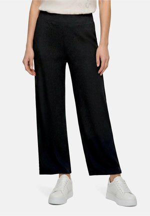 Femme portant un pantalon noir à jambes larges et des baskets blanches, debout, les mains le long du corps, sur un fond uni.