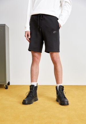 Mann trägt schwarze Nike-Shorts, weiße Socken, schwarze Schnürstiefel und ein weißes Langarmshirt und steht auf einem gelben Teppich.