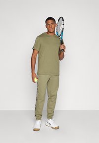 Ropa deportiva de color verde oliva con una camiseta de manga corta y pantalones de chándal a juego. Presenta un logo en la camiseta y rayas laterales en los pantalones. Sosteniendo una raqueta de tenis y una pelota.