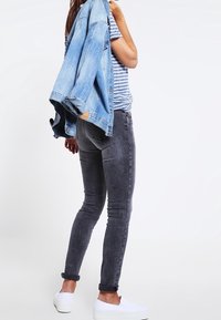 Jeans ajustados grises con una textura desgastada, combinados con una camisa a rayas. Una chaqueta de mezclilla clara está colgada sobre el hombro. Se llevan zapatillas blancas.