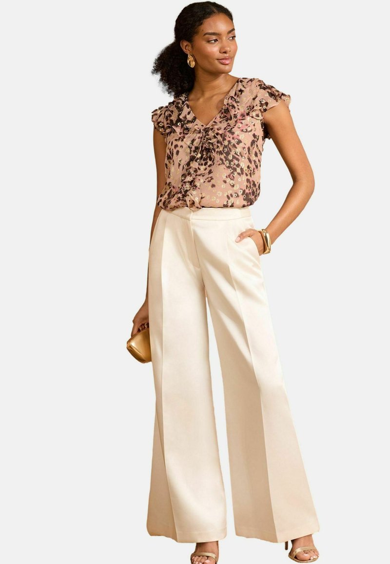Blouse with floral pattern in beige and black, v-neck, ruffled sleeves; paired with wide-leg cream pants; gold accessories.
