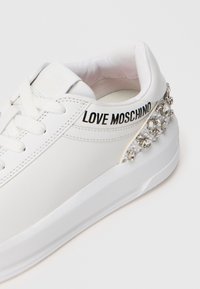 Bijele kožne tenisice s glatkom površinom, s crnim logotipom, vezicama i ukrašenim rhinestones-ima na peti.