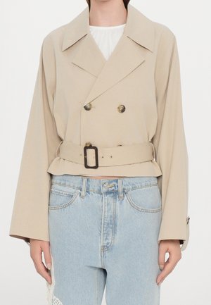 Beige lyhyt kaksirivinen takki, jossa leveä kaulus ja vyö, yhdistetty vaaleansinisiin korkeavyötäröisiin farkkuihin, joissa on pitsikoristelu toisessa lahkeessa.