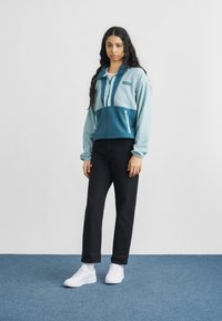 Ζακέτα φλις με φερμουάρ σε ανοιχτό μπλε και τιρκουάζ, με δύο μπροστινές τσέπες, ριγέ μανσέτες και λευκό μπλουζάκι από κάτω. Μαύρο παντελόνι και λευκά παπούτσια.