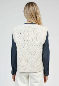 Gilet en tricot crème avec un motif en relief, doté d'un large col et d'emmanchures, porté par-dessus une chemise en denim noir.
