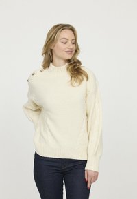 Pull en tricot couleur crème avec des motifs en câble, des poignets côtelés et un col montant. Présente des boutons décoratifs sur l'épaule gauche.