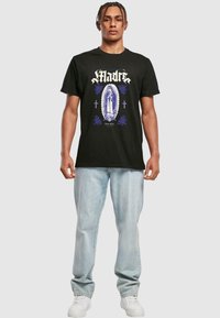Zwart katoen T-shirt met een centraal graphic van een heilige, verfraaid met blauwe bloemen- en kruisontwerpen. Gecombineerd met lichtblauwe jeans en witte sneakers.