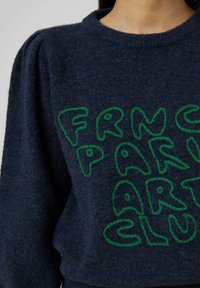 Pull en maille bleu marine avec texte vert brodé disant "FRANCE PARIS ART CLUB." Comprend des manches bouffantes et un col rond.